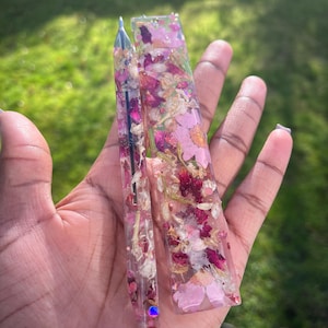 May include: Two clear resin accessories, one pen-shaped and one rectangular, both filled with dried pink and red flowers. The pen has a silver tip. The items are held in a hand against a blurred green background.