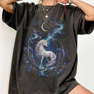 Women’s fantasy graphic t-shirt featuring a  white unicorn standing in an enchanted celestial forest surrounded by glowing moonlight, magical stars, mystical trees, cosmic details, and dreamy fantasy artwork. The design has a vintage-inspired magical aesthetic with fairytale atmosphere, celestial night elements, flowing fantasy illustration, and enchanted woodland scenery printed on a relaxed fit graphic tee for unicorn lovers, fairycore fashion fans, and mystical fantasy style outfits.
