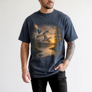 Vintage-inspired wildlife t-shirt featuring realistic flying mallard ducks over a mountain lake at sunrise with retro nature artwork, natural islands, and rustic outdoor aesthetic. Comfort Colors 1717 garment-dyed t-shirt