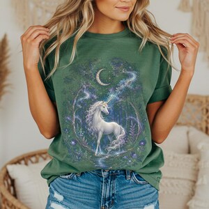 Women’s fantasy graphic t-shirt featuring a  white unicorn standing in an enchanted celestial forest surrounded by glowing moonlight, magical stars, mystical trees, cosmic details, and dreamy fantasy artwork. The design has a vintage-inspired magical aesthetic with fairytale atmosphere, celestial night elements, flowing fantasy illustration, and enchanted woodland scenery printed on a relaxed fit graphic tee for unicorn lovers, fairycore fashion fans, and mystical fantasy style outfits.