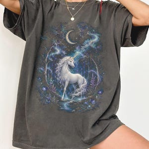 Women’s fantasy graphic t-shirt featuring a  white unicorn standing in an enchanted celestial forest surrounded by glowing moonlight, magical stars, mystical trees, cosmic details, and dreamy fantasy artwork. The design has a vintage-inspired magical aesthetic with fairytale atmosphere, celestial night elements, flowing fantasy illustration, and enchanted woodland scenery printed on a relaxed fit graphic tee for unicorn lovers, fairycore fashion fans, and mystical fantasy style outfits.