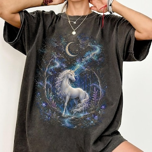 Women’s fantasy graphic t-shirt featuring a  white unicorn standing in an enchanted celestial forest surrounded by glowing moonlight, magical stars, mystical trees, cosmic details, and dreamy fantasy artwork. The design has a vintage-inspired magical aesthetic with fairytale atmosphere, celestial night elements, flowing fantasy illustration, and enchanted woodland scenery printed on a relaxed fit graphic tee for unicorn lovers, fairycore fashion fans, and mystical fantasy style outfits.