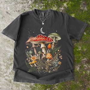 May include: A charcoal gray short-sleeve t-shirt featuring a graphic print of various mushrooms, ferns, and a crescent moon. A silver-toned necklace with a small star-shaped pendant rests on the neckline of the shirt, centered above the graphic.