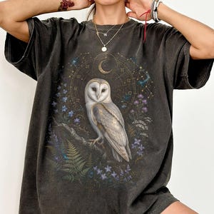 Może przedstawiać: Ciemnoszary t-shirt z krótkim rękawem z centralną grafiką przedstawiającą płomykówkę siedzącą na gałęzi. Wzór zawiera półksiężyc, znaki zodiaku, paprocie oraz fioletowe i niebieskie polne kwiaty otaczające sowę.
