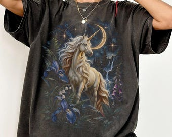 May include: A charcoal gray short-sleeve t-shirt featuring a graphic print of a white unicorn with a flowing mane standing in a forest. The design includes a crescent moon, stars, a waterfall, and dark blue iris and lily of the valley flowers.