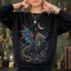 Puede incluir: Una sudadera negra de manga larga con un estampado gráfico de un dragón con alas azules y doradas, una luna creciente y elementos celestiales estrellados. El diseño incluye líneas doradas intrincadas y un castillo.