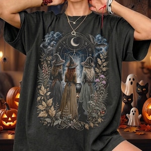 Può includere: Una t-shirt grigio antracite con una stampa grafica di tre figure con cappelli a punta in piedi davanti a un calderone. Il design include una luna crescente, simboli celesti, un corvo, un pipistrello e fogliame. Ha maniche corte e girocollo.