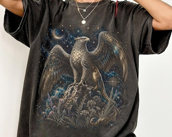 May include: A charcoal gray short-sleeve t-shirt featuring a detailed graphic print of a golden griffin perched on a rocky cliff. The background of the illustration depicts a dark blue starry night sky with a crescent moon and celestial elements.