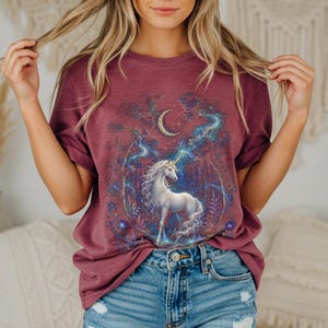 Women’s fantasy graphic t-shirt featuring a  white unicorn standing in an enchanted celestial forest surrounded by glowing moonlight, magical stars, mystical trees, cosmic details, and dreamy fantasy artwork. The design has a vintage-inspired magical aesthetic with fairytale atmosphere, celestial night elements, flowing fantasy illustration, and enchanted woodland scenery printed on a relaxed fit graphic tee for unicorn lovers, fairycore fashion fans, and mystical fantasy style outfits.