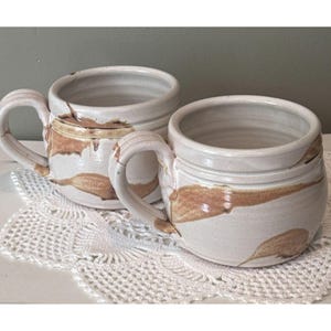 May include: Two handmade ceramic mugs with a rounded shape and a wide opening. The mugs feature a light cream-colored glaze with irregular, horizontal brown brushstroke patterns and a ribbed texture near the top rim, each with a curved handle.