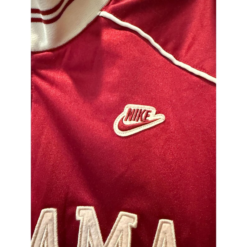 May include: A red athletic jacket with a white collar and trim. The Nike logo is embroidered on the chest. The jacket has white lettering at the bottom. The fabric appears to be a soft, velvety material.