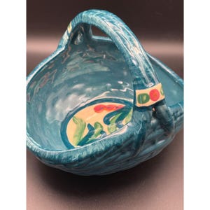 May include: A turquoise ceramic basket with a handle. The basket has a textured exterior and a painted oval design with red and green floral elements. The handle has decorative accents.