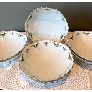May include: A set of four white porcelain bowls with scalloped edges and a delicate green and silver floral pattern along the rims. The bowls feature a smooth, glossy finish and a rounded shape, arranged in a cluster to display the decorative details.