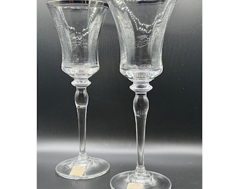 May include: Two clear crystal wine glasses with silver rims. The glasses have a detailed etched design and long stems. The glasses are on a dark background.