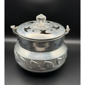 May include: A silver-colored incense burner with a lid and a clear, faceted knob. The lid has decorative cutouts. The body of the burner features a leaf design. The handles are gold-colored. The incense burner is on a black background.