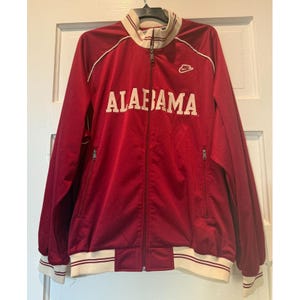 May include: A crimson red zip-up track jacket with the word "ALABAMA" in white block letters across the chest. The jacket has a white collar, cuffs, and hem with red stripes. A small Nike logo is on the upper right chest.