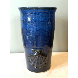 May include: A tall, cylindrical, cobalt blue ceramic vase with a glossy finish. The vase features a raised seashell design near the base. The rim and seashell details are a darker shade of blue.