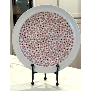 May include: A white ceramic plate with a floral pattern in shades of pink and red. The plate is displayed on a black metal stand. The floral design covers the majority of the plate's surface, creating a decorative piece.
