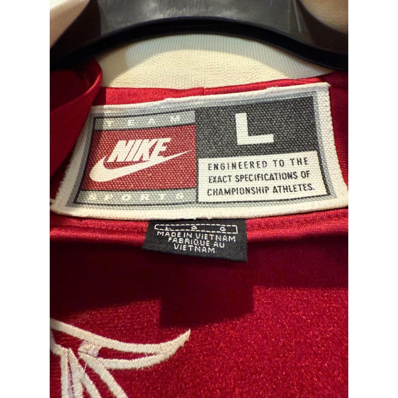 May include: Close-up of a red Nike sports jacket tag. The tag features the Nike logo, the letter "L", and text that reads "Engineered to the exact specifications of championship athletes." The tag also indicates the jacket was made in Vietnam.