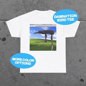 Op de afbeelding: Wit T-shirt met een afbeelding van een vogel met lange poten tegen een groene achtergrond en blauwe lucht. Het shirt heeft de tekst "DAMN!!!!!!!!!!!!" boven de vogel en "DAMNATION BIRD TEE" in een blauwe ovaal.