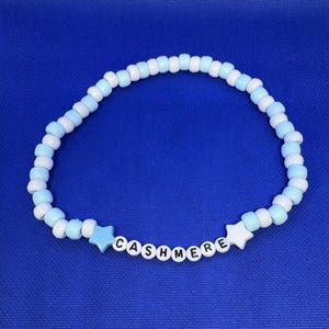 May include: A beaded bracelet with light blue and white beads. The word "CASHMERE" is spelled out in white letter beads. Two star-shaped beads, one light blue and one white, flank the word. The bracelet is on a blue background.