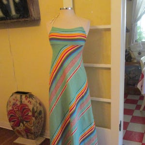 May include: A light green halter dress with colorful horizontal stripes across the bust and diagonal stripes down the skirt. The dress is displayed on a mannequin. The dress is a maxi length.