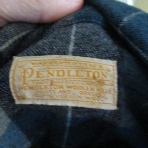 Puede incluir: Primer plano de una etiqueta Pendleton cosida en una tela de cuadros azul y negro. La etiqueta es rectangular con fondo crema y letras doradas. El texto dice "WARRANTED TO BE A PENDLETON" y otros detalles del producto.