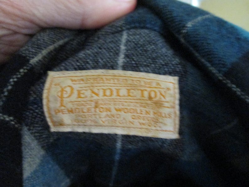 Puede incluir: Primer plano de una etiqueta Pendleton cosida en una tela de cuadros azul y negro. La etiqueta es rectangular con fondo crema y letras doradas. El texto dice "WARRANTED TO BE A PENDLETON" y otros detalles del producto.