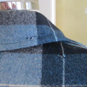Puede incluir: Primer plano de una tela a cuadros azul, negro y gris. La tela est&aacute; tejida con un aspecto texturizado. El cuello de una prenda es visible, con una costura y una peque&ntilde;a imperfecci&oacute;n.