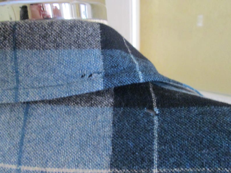 Puede incluir: Primer plano de una tela a cuadros azul, negro y gris. La tela est&aacute; tejida con un aspecto texturizado. El cuello de una prenda es visible, con una costura y una peque&ntilde;a imperfecci&oacute;n.