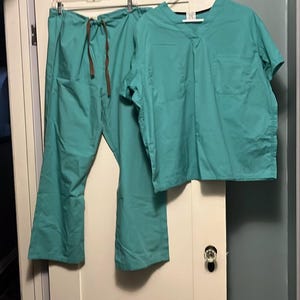 May include: Teal-colored medical scrubs set, including a short-sleeved top and pants with a drawstring waist. The scrubs are hanging on a white hanger against a white door and a light blue wall.