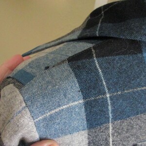 Puede incluir: Primer plano de una camisa a cuadros azul, negra y gris. Se ve el cuello de la camisa, junto con el hombro y la parte superior de la manga. El estampado de cuadros presenta una cuadr&iacute;cula de l&iacute;neas blancas.