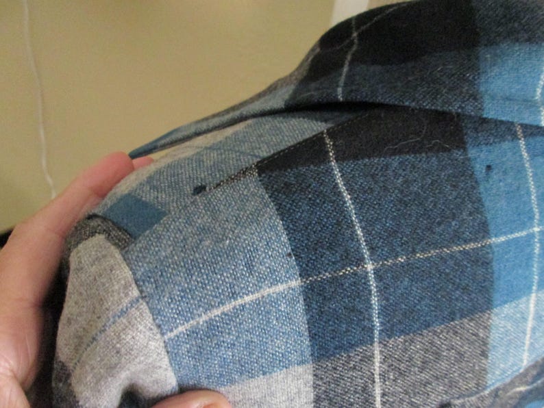 Puede incluir: Primer plano de una camisa a cuadros azul, negra y gris. Se ve el cuello de la camisa, junto con el hombro y la parte superior de la manga. El estampado de cuadros presenta una cuadr&iacute;cula de l&iacute;neas blancas.