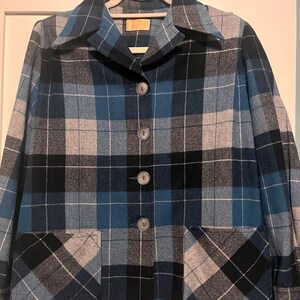 Puede incluir: Chaqueta vintage a cuadros en tonos azules, negros y grises, con un dise&ntilde;o cl&aacute;sico abotonado y dos bolsillos delanteros. La chaqueta est&aacute; colgada en una percha blanca.