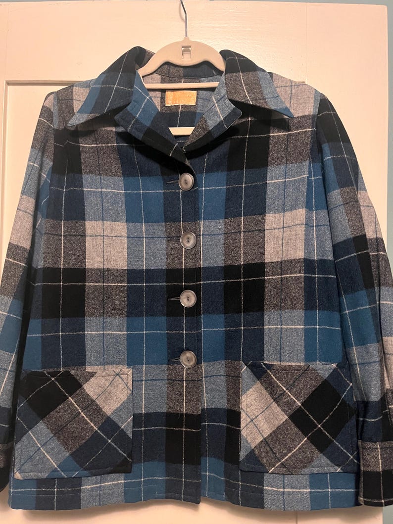 Puede incluir: Chaqueta vintage a cuadros en tonos azules, negros y grises, con un dise&ntilde;o cl&aacute;sico abotonado y dos bolsillos delanteros. La chaqueta est&aacute; colgada en una percha blanca.