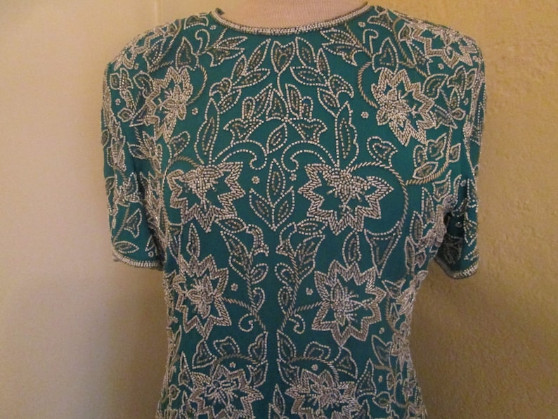 May include: A teal blue top with a silver floral pattern. The top is made of a soft, silky fabric and is fully beaded.