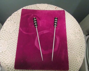 May include: Two silver-toned hat pins with black and silver beaded tops rest on a deep magenta velvet surface. The pins are displayed on a white lace doily, creating a vintage aesthetic. The pins are designed for securing hats.