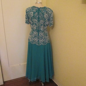 May include: A teal formal dress with a white beaded bodice and a flowing skirt. The dress is on a mannequin and is displayed against a light yellow wall.
