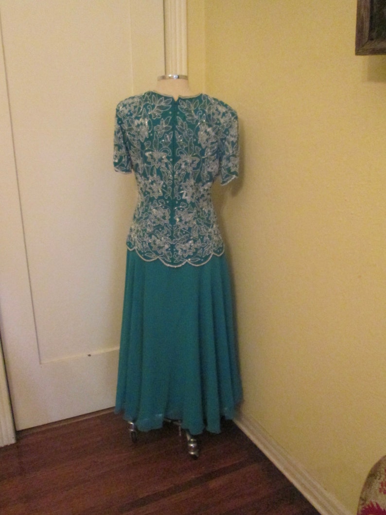 May include: A teal formal dress with a white beaded bodice and a flowing skirt. The dress is on a mannequin and is displayed against a light yellow wall.