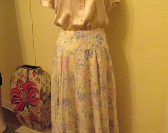 May include: A light beige satin blouse with a collared neckline and short sleeves paired with a long, flowing floral skirt. The skirt has a light yellow background with pink, blue, and white floral patterns.
