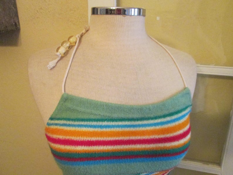 May include: A colorful halter top with horizontal stripes in shades of teal, orange, pink, blue, and white. The top is displayed on a mannequin and features a beaded strap and a tassel.