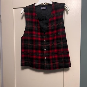 May include: A sleeveless vest featuring a red and black plaid pattern. The garment has a V-neckline, a black interior lining, and a front button closure with three metallic buttons. Two horizontal welt pockets are positioned at the lower front.