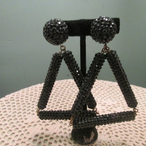 Pode incluir: Um par de brincos de strass preto com formato triangular. Os brincos são feitos de várias barras conectadas cobertas de strass.