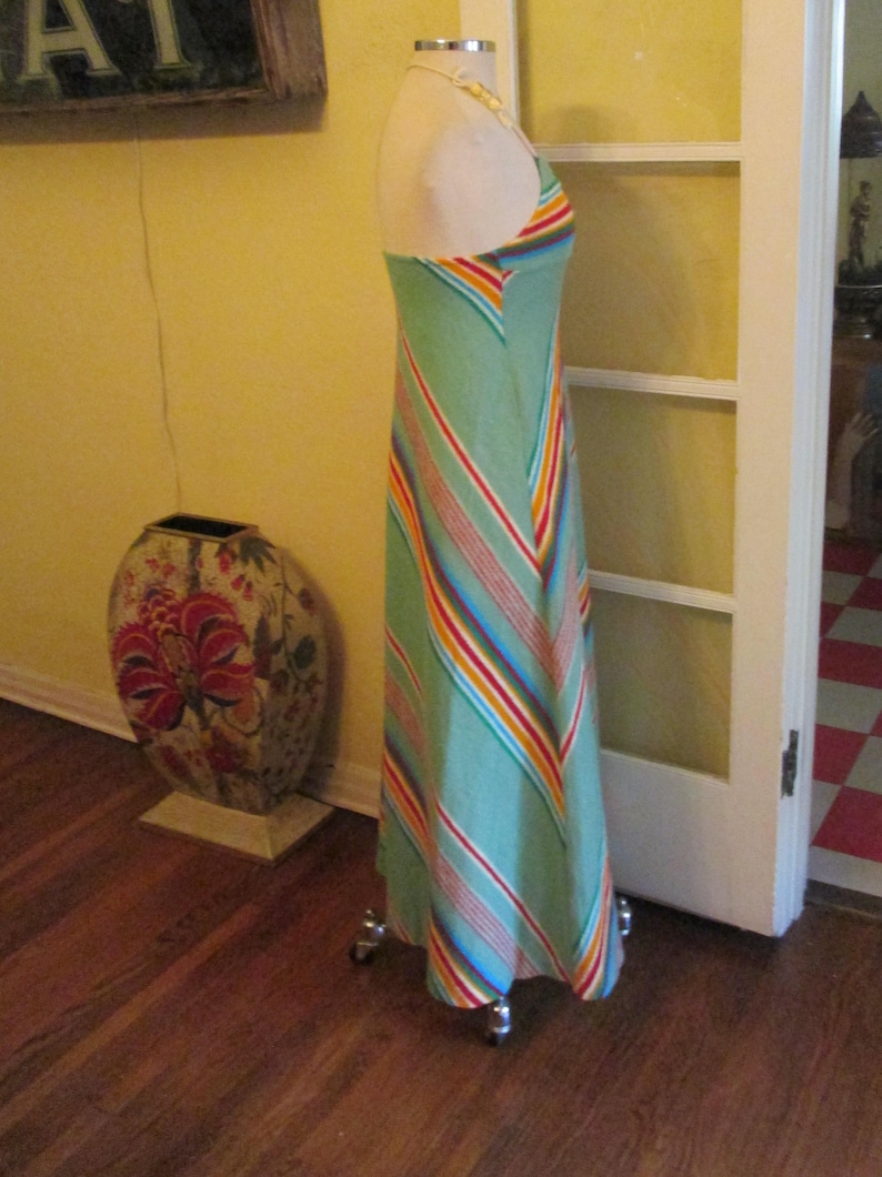 May include: A light green halter maxi dress with colorful diagonal stripes. The dress is displayed on a mannequin. The stripes include red, orange, yellow, and blue. The dress is floor length.
