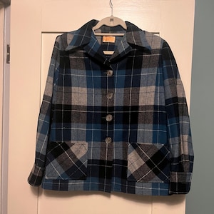 Puede incluir: Una chaqueta a cuadros azul, negro y gris con dos bolsillos delanteros y tres botones. La chaqueta est&aacute; colgada de una percha blanca contra una puerta blanca.