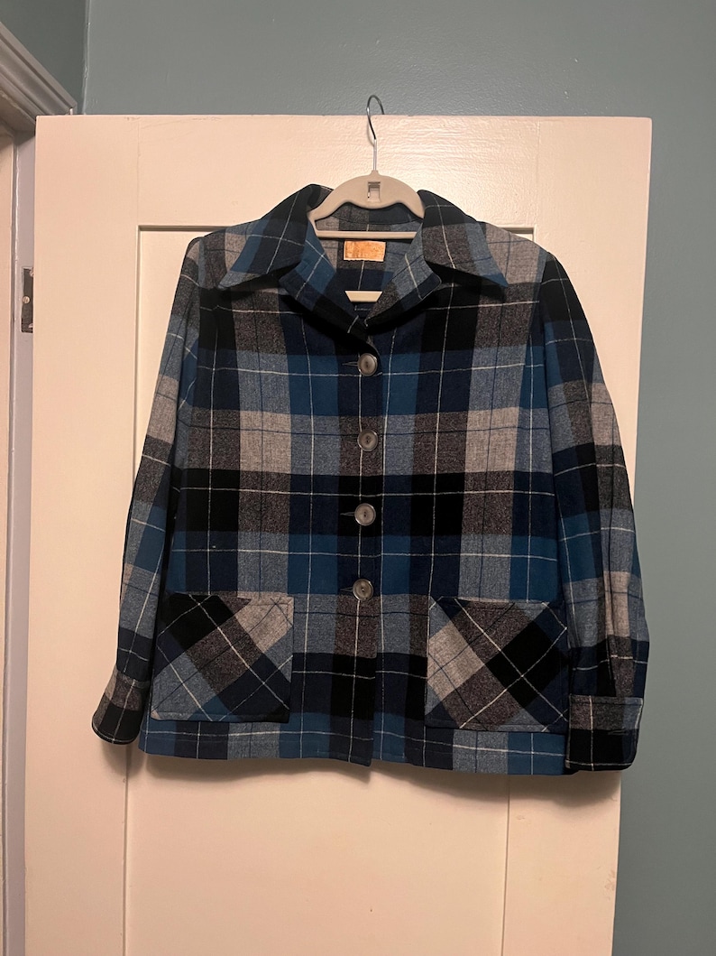 Puede incluir: Una chaqueta a cuadros azul, negro y gris con dos bolsillos delanteros y tres botones. La chaqueta est&aacute; colgada de una percha blanca contra una puerta blanca.