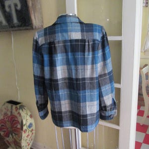 Puede incluir: Una camisa a cuadros azul, negra y gris sobre un maniqu&iacute;. La camisa de manga larga tiene una parte delantera y pu&ntilde;os abotonados. La parte trasera de la camisa es visible, mostrando el cuello y el patr&oacute;n de cuadros. La camisa se exhibe frente a una puerta blanca.