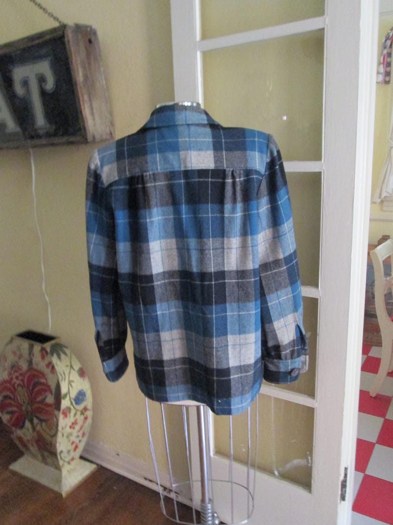 Puede incluir: Una camisa a cuadros azul, negra y gris sobre un maniqu&iacute;. La camisa de manga larga tiene una parte delantera y pu&ntilde;os abotonados. La parte trasera de la camisa es visible, mostrando el cuello y el patr&oacute;n de cuadros. La camisa se exhibe frente a una puerta blanca.