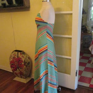 May include: A vintage halter-neck maxi dress on a mannequin. The dress features a light green background with colorful horizontal stripes in red, orange, yellow, blue, and white. The dress is floor-length and has a flowing silhouette, perfect for a retro look.
