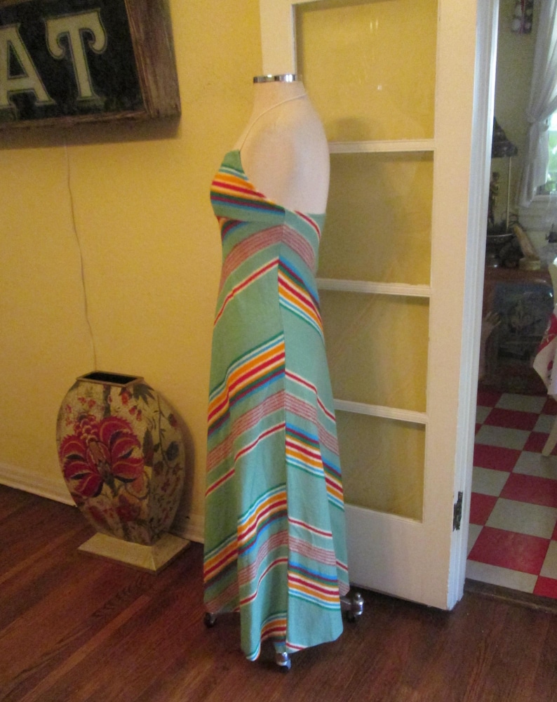 May include: A vintage halter-neck maxi dress on a mannequin. The dress features a light green background with colorful horizontal stripes in red, orange, yellow, blue, and white. The dress is floor-length and has a flowing silhouette, perfect for a retro look.